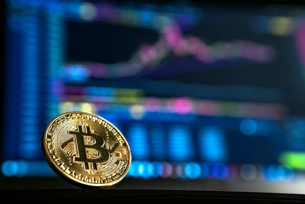A physical Bitcoin illustration in front of a market screen